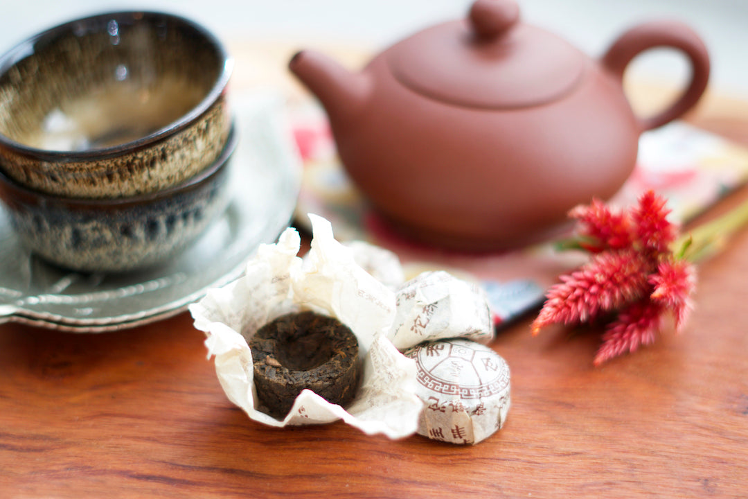 foreground: unwrapped and wrapped tuocha or small nest of aged pu erh tea. Background: small cups and a red yixing clay teapot