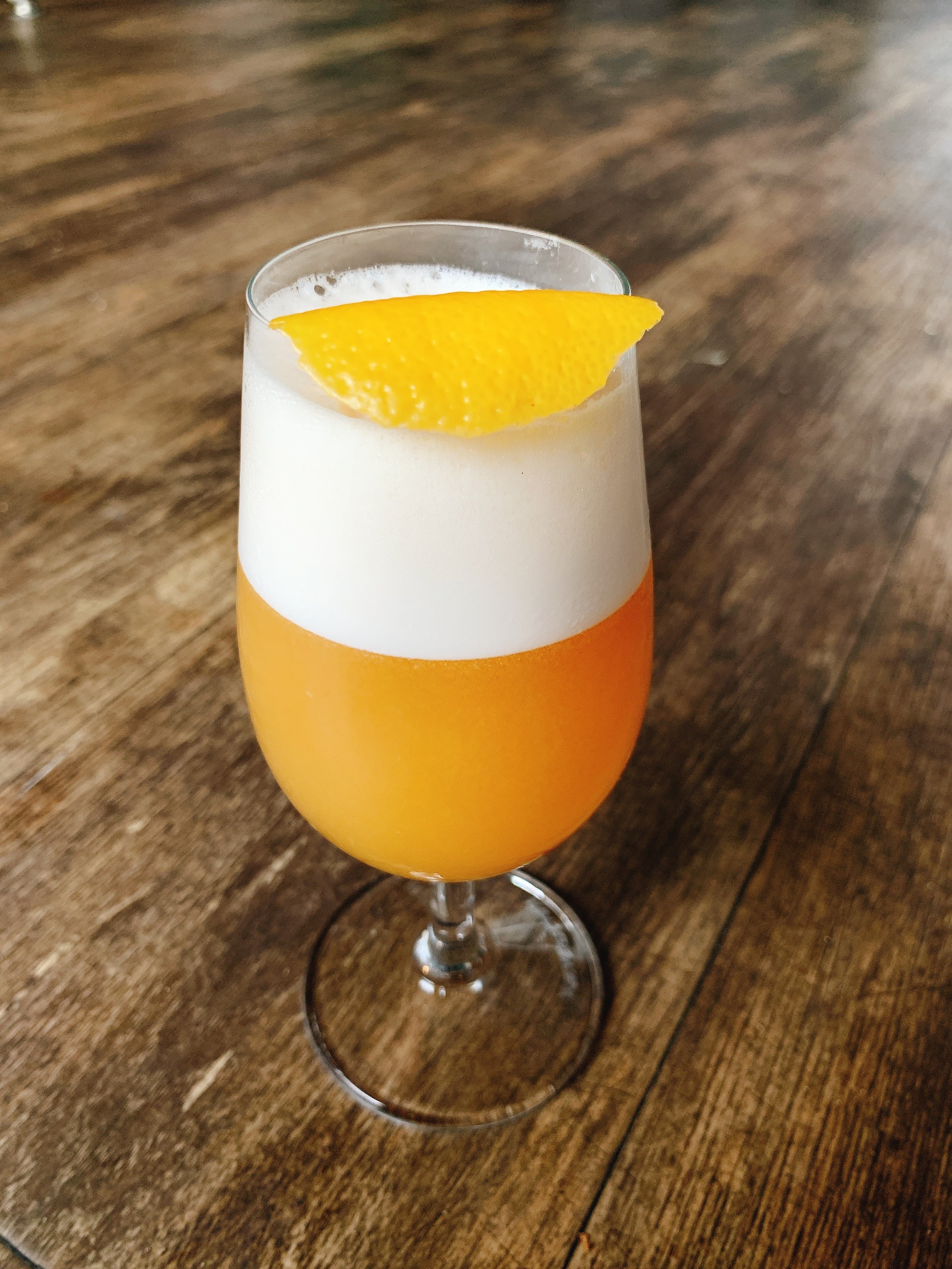 small stemmed cocktail glass on a wooden bartop with a orange cocktail topped with egg white foam and orange peel