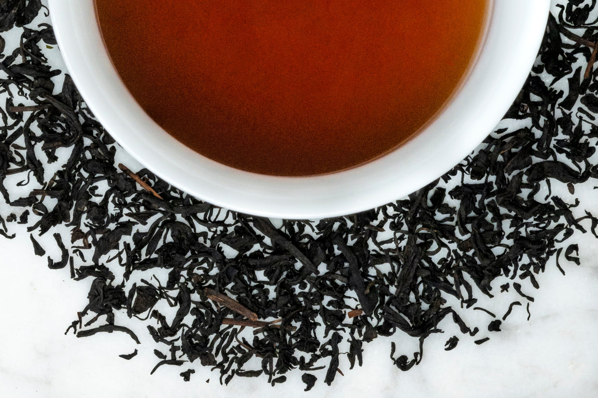 2020 Jeju leaves and brewed tea. Dark brown, almost black twisted leaves of varying sizes and shapes surround a cup of dark reddish-brown brewed pu erh tea.