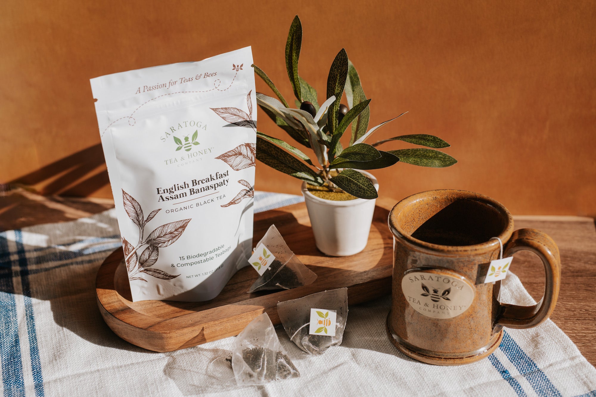 Aesthetic scene of pyramid tea sachets with bee tags strewn next to a tan stoneware logo mug from Saratoga Tea & Honey Co. The scene also includes the sachet packaging pouch and some greenery.