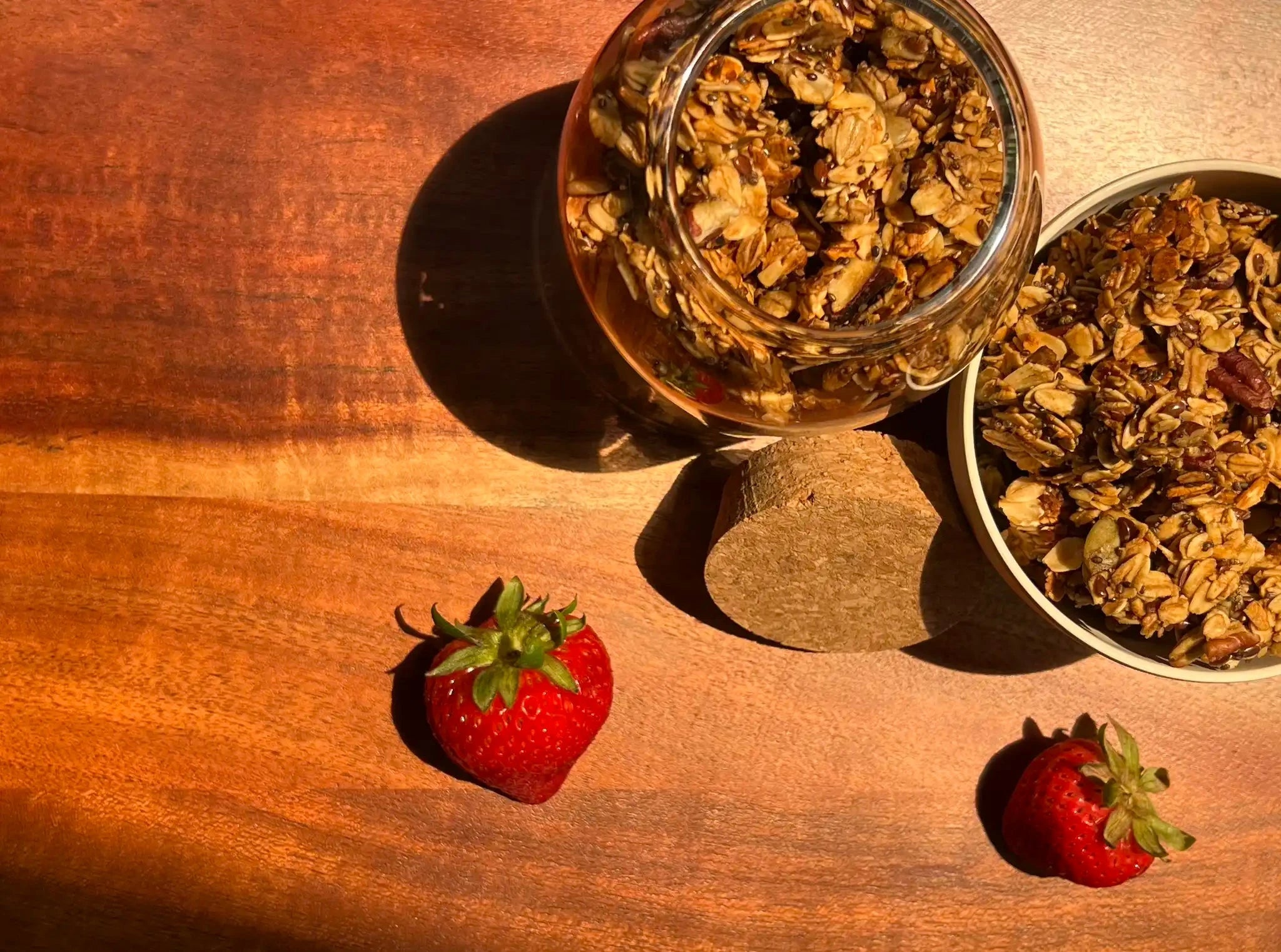 aesthetic image of homemade granola in a glass jar and white bowl on a wood surface with scattered strawberries