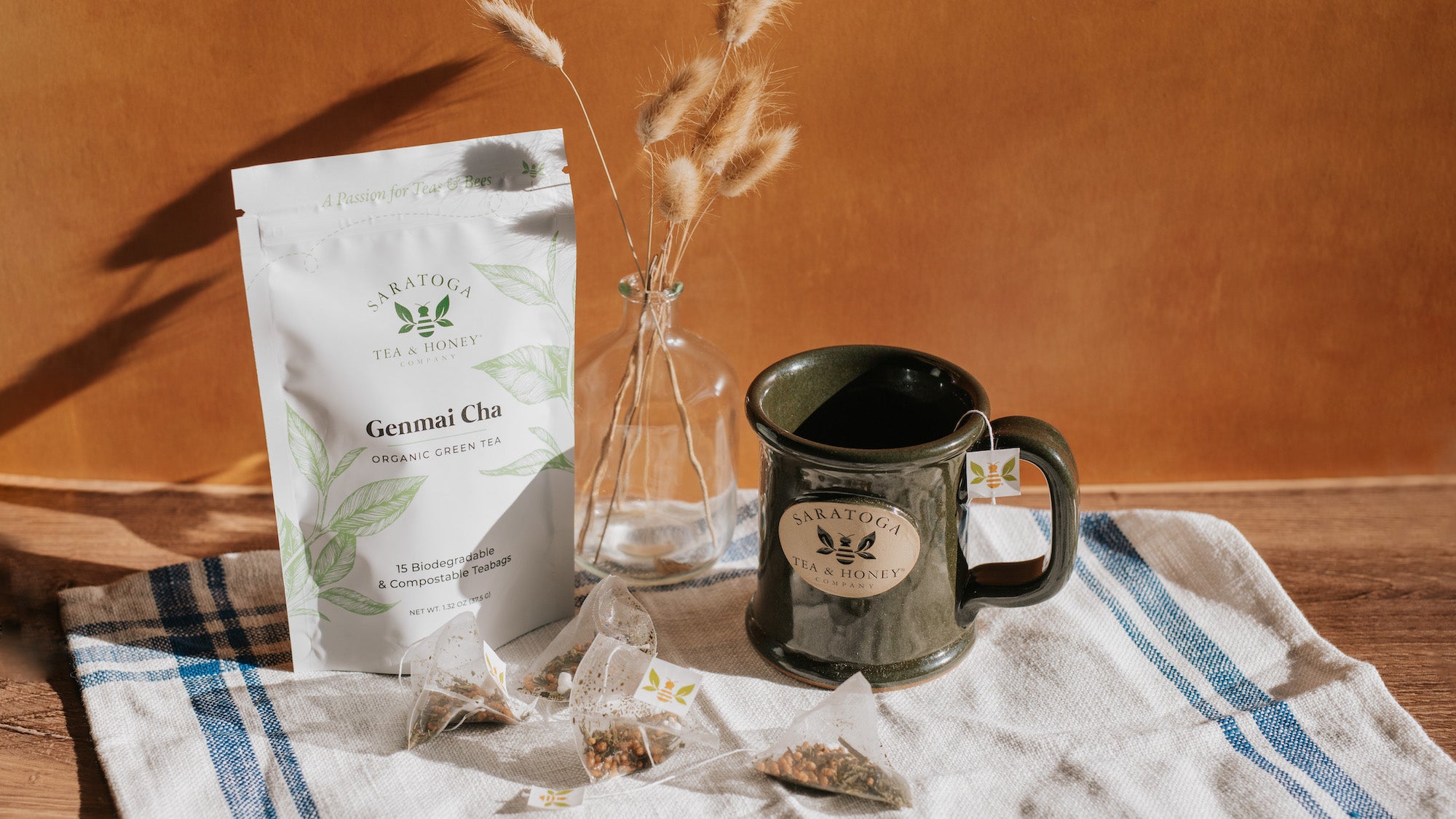Aesthetic scene of Genmai Cha green tea sachets alongside a dark green stoneware logo mug from Saratoga Tea & Honey Co. The sachets are strewn in front of their pouch and the mug has a bee tag on a string hanging over the side.