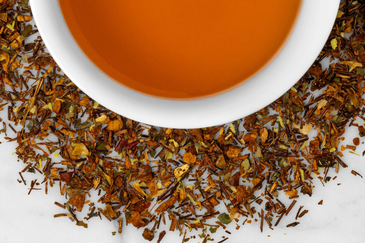Close up of dry and brewed Island Glow. The dry tea is composed of rooibos with turmeric, mango, and coconut pieces surrounding a cup of brewed, orange-hued tea.