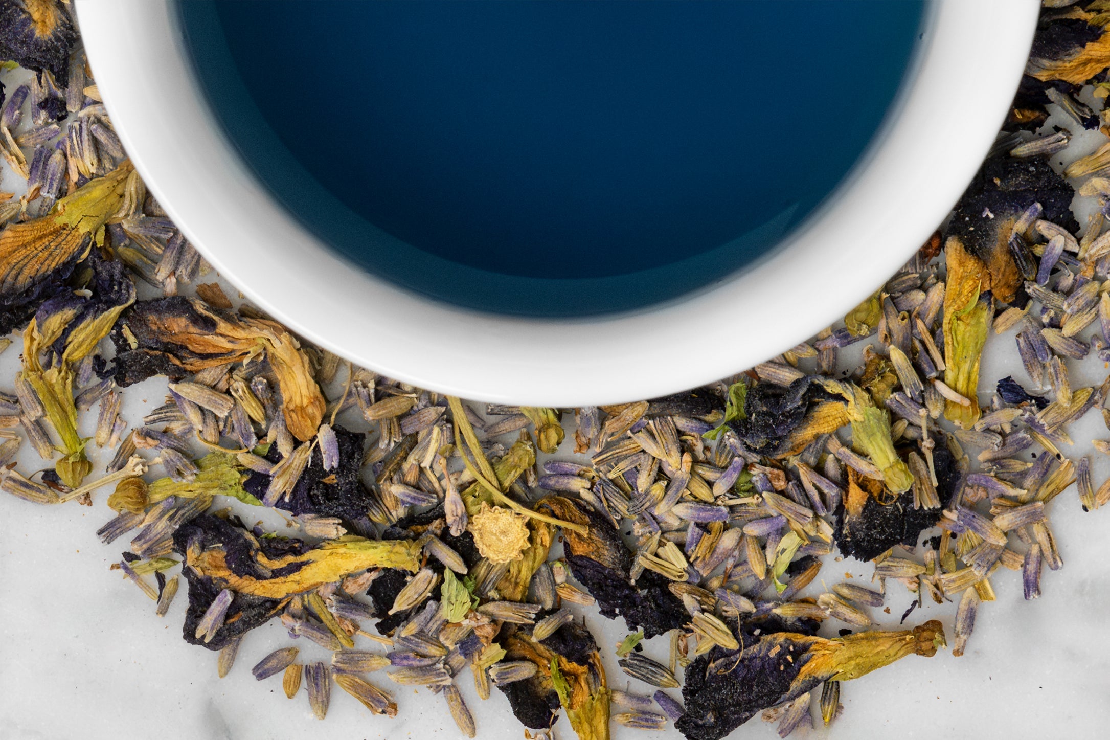 Close up of dry and brewed Karner Blue Lavender. The dry tea is composed of lavender buds and butterfly pea flower blossoms surrounding a cup of brewed, deep blue tea.