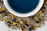 Close up of dry and brewed Karner Blue Lavender. The dry tea is composed of lavender buds and butterfly pea flower blossoms surrounding a cup of brewed, deep blue tea.