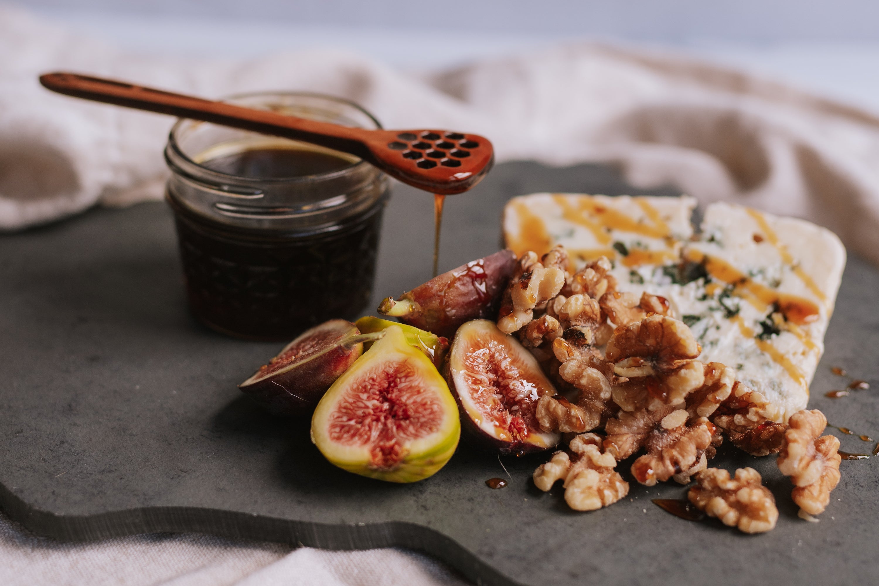 dark and rich honey in a glass jar and dripping off a wooden dipper and on to a cheese board with bleu cheese, fig, and walnuts