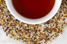Brewed and dry Nana's Pumpkin Pie herbal tisane. A dark, reddish-brown cup of brewed tea is surrounded by small loose leaf rooibos, ginger, honeybush, clove, and nutmeg. 