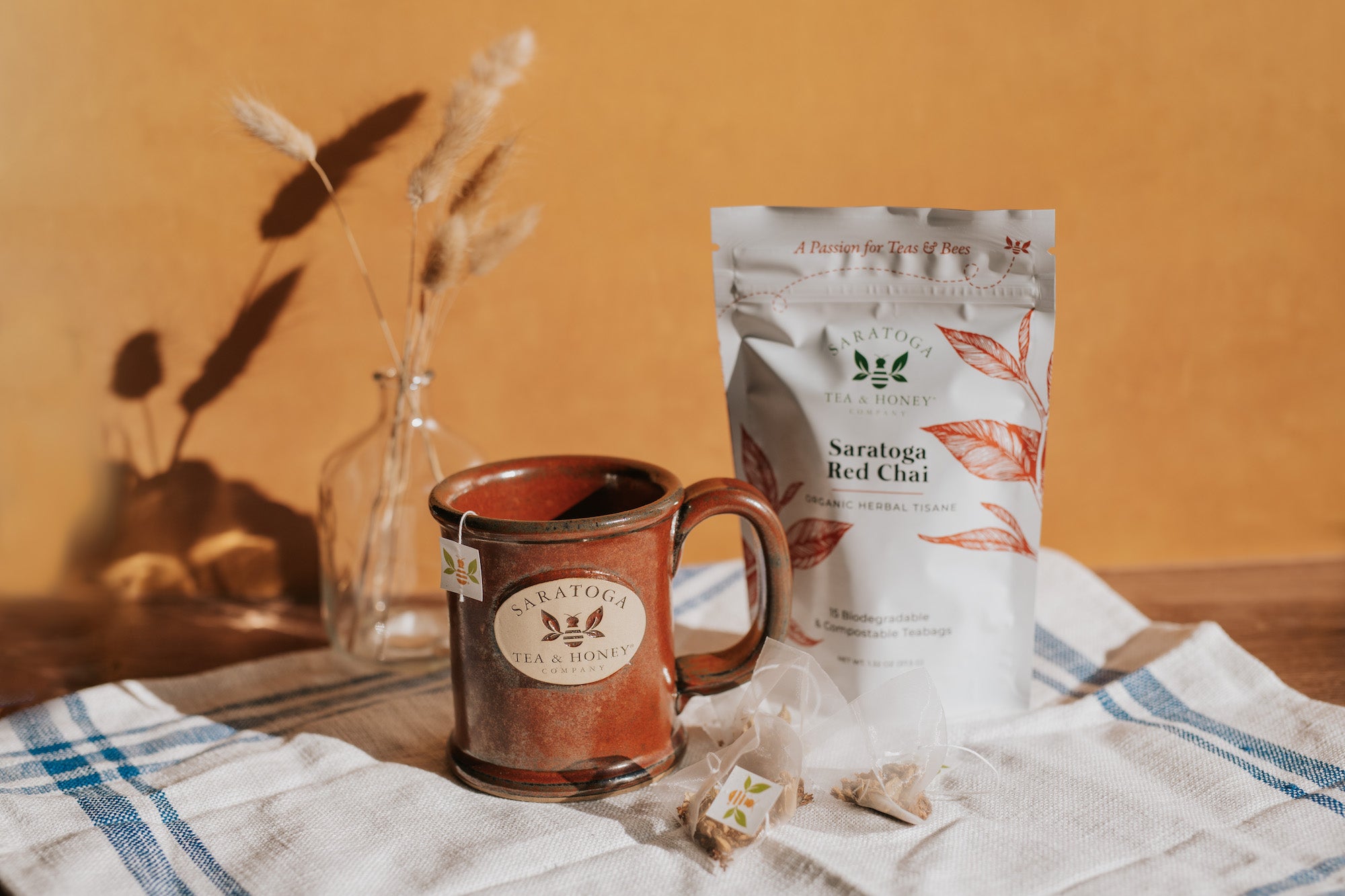 Aesthetic scene of a pouch of Saratoga Red Chai tea bags alongside a Saratoga Tea & Honey Co. stoneware mug and a small pile of pyramid tea bags with bee tags.