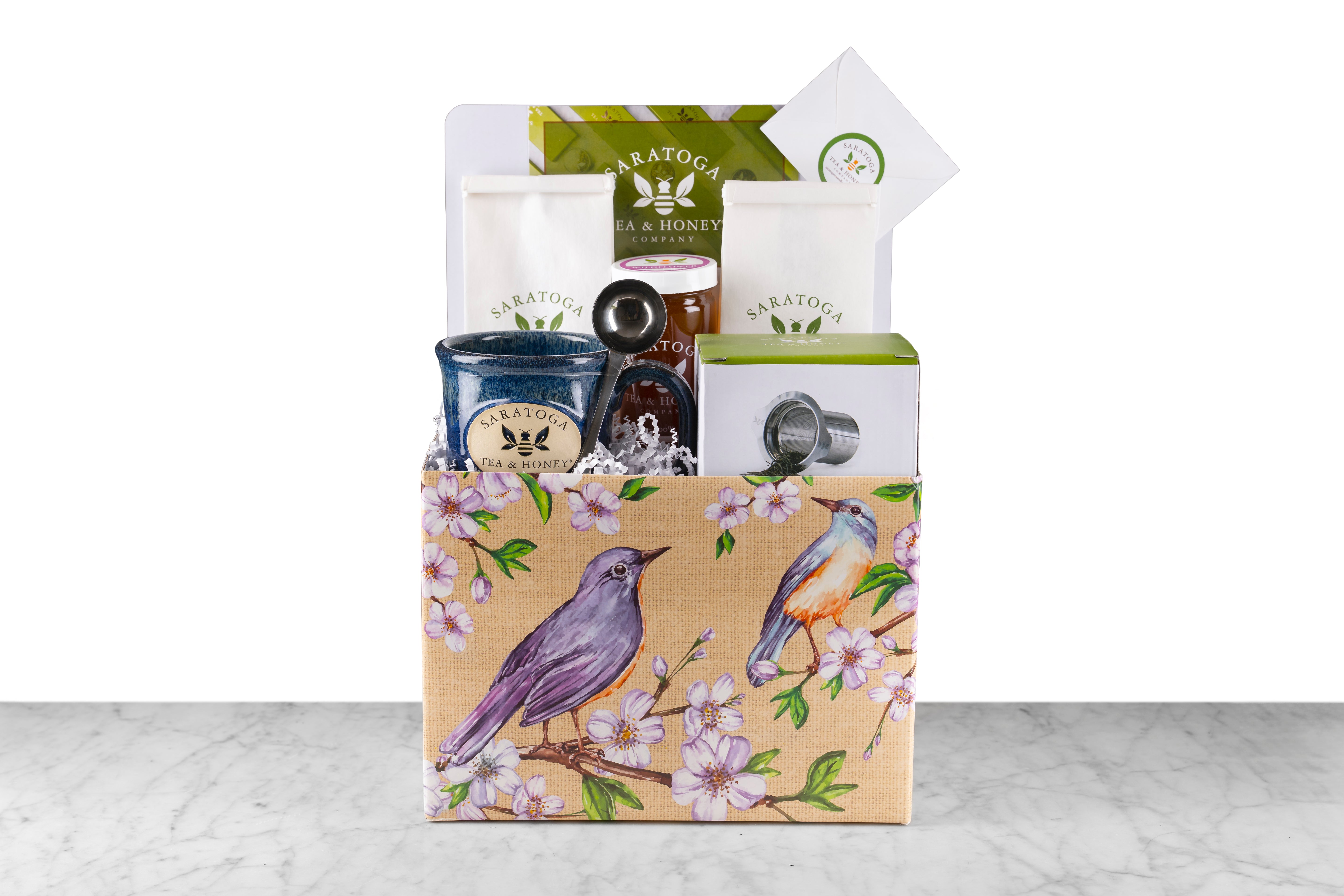 A gift basket containing a stoneware mug with a bee logo, a stainless steel tea infuser, a stainless steel tea scoop, two bags of loose leaf tea, a jar of wildflower honey, and a gift card.