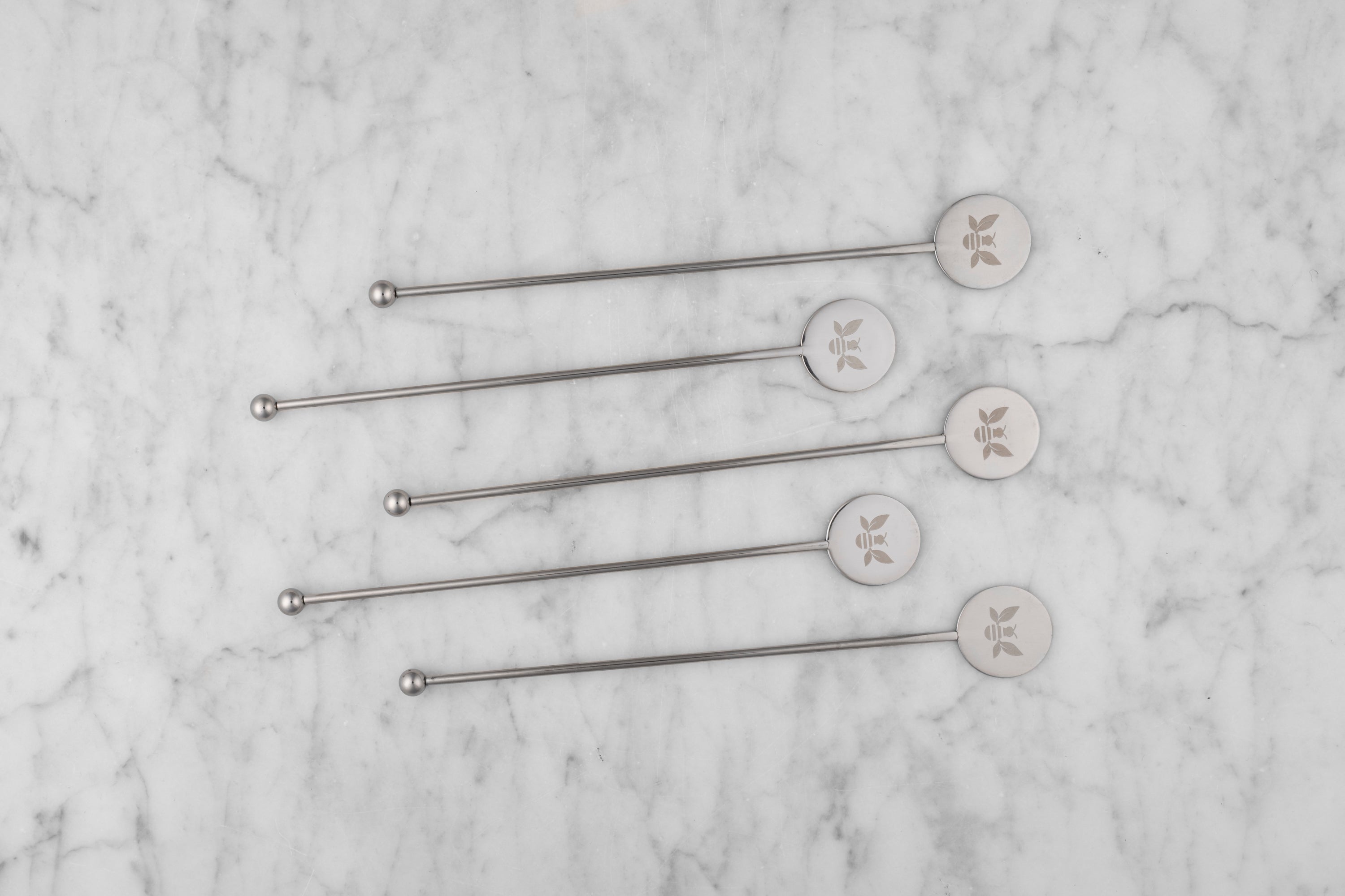 silver cocktail stirrers with etched Saratoga Tea & Honey Co. bee lined up on a marble background