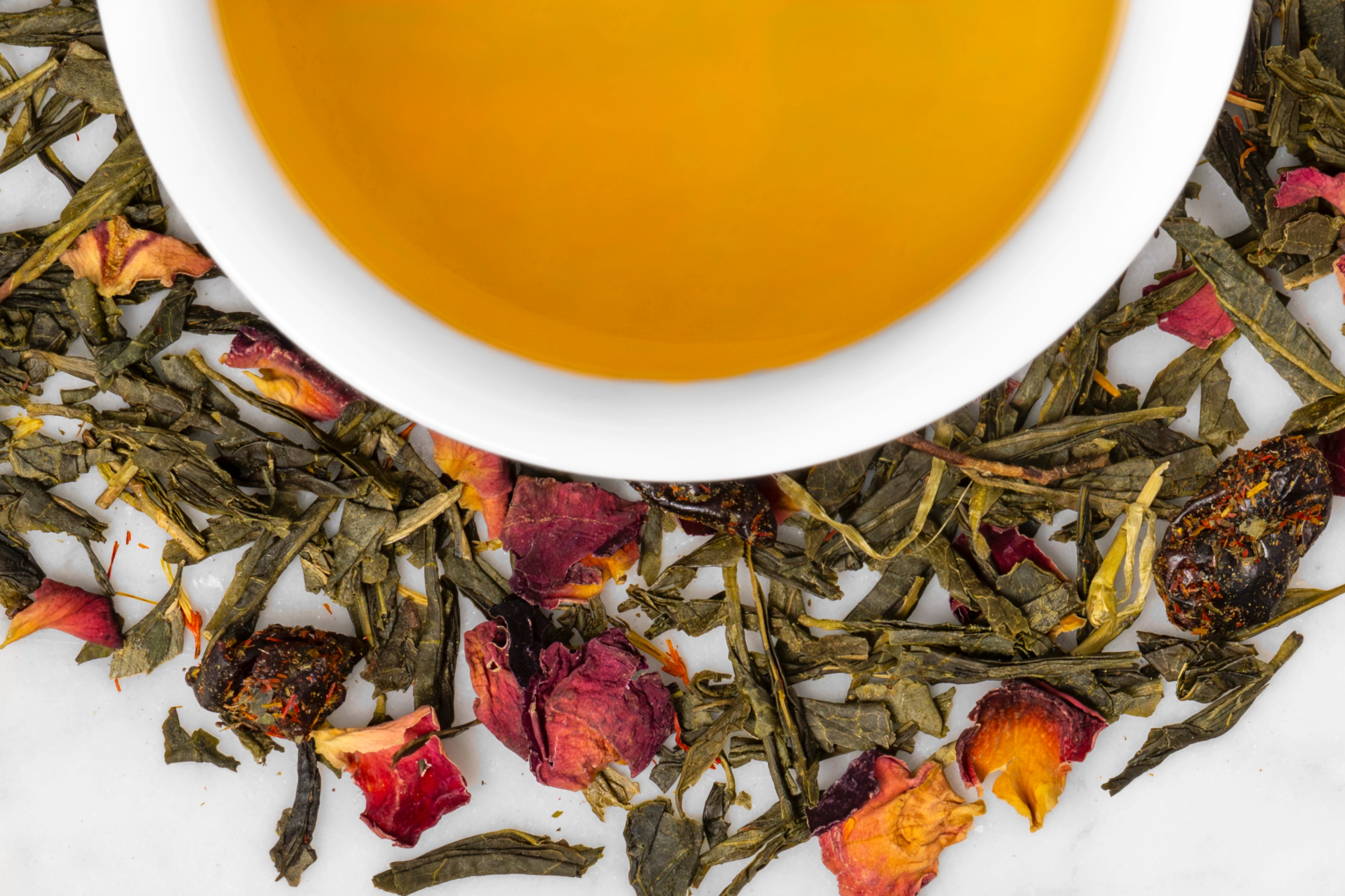 Dried green tea leaves and rose petals surrounding a cup of Summer Fruit tea.