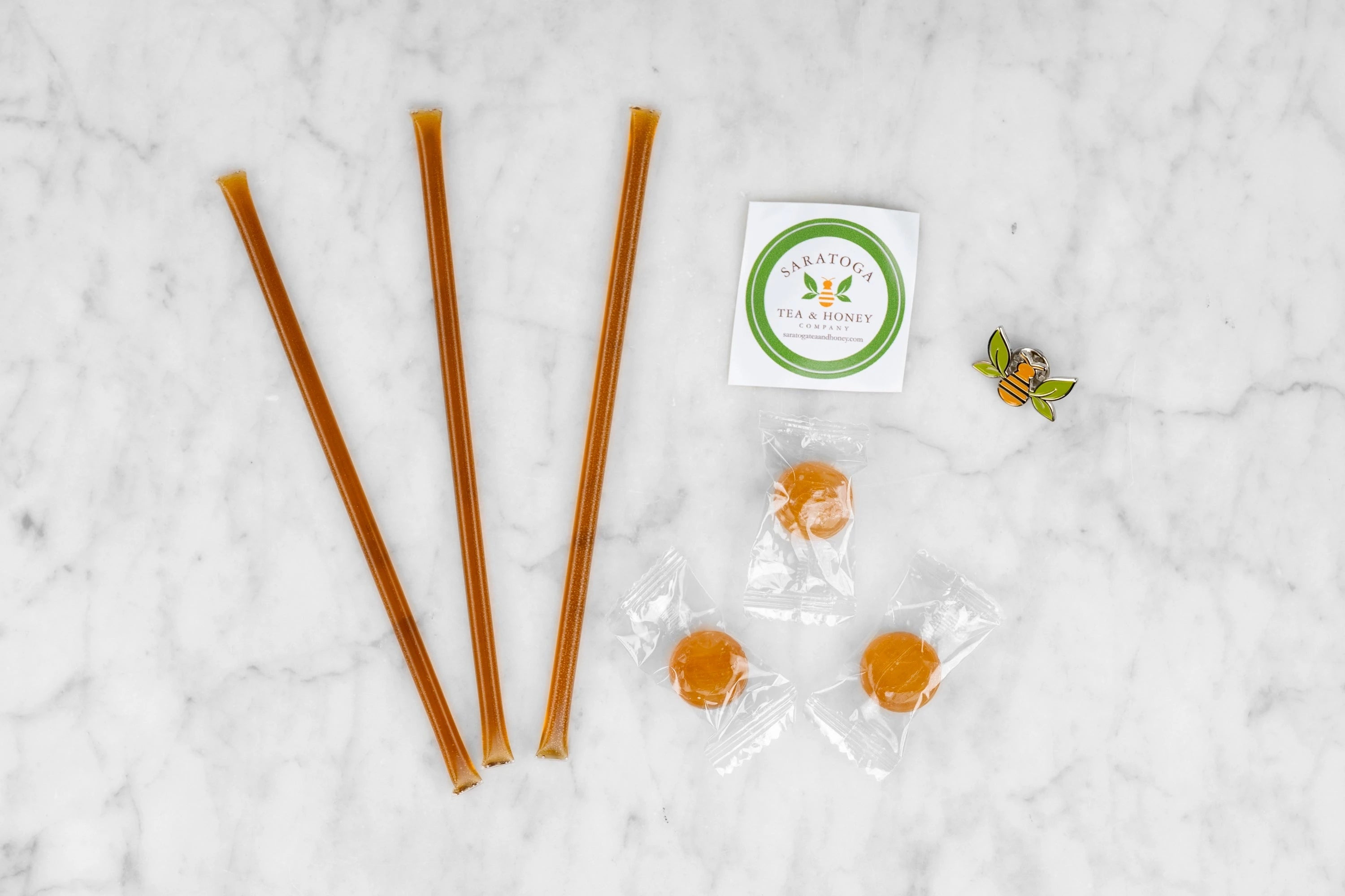 The Swag Bag contents: three honey sticks, three round hard honey candies, a Saratoga Tea & Honey Co. sticker and a tea bee enamel pin.