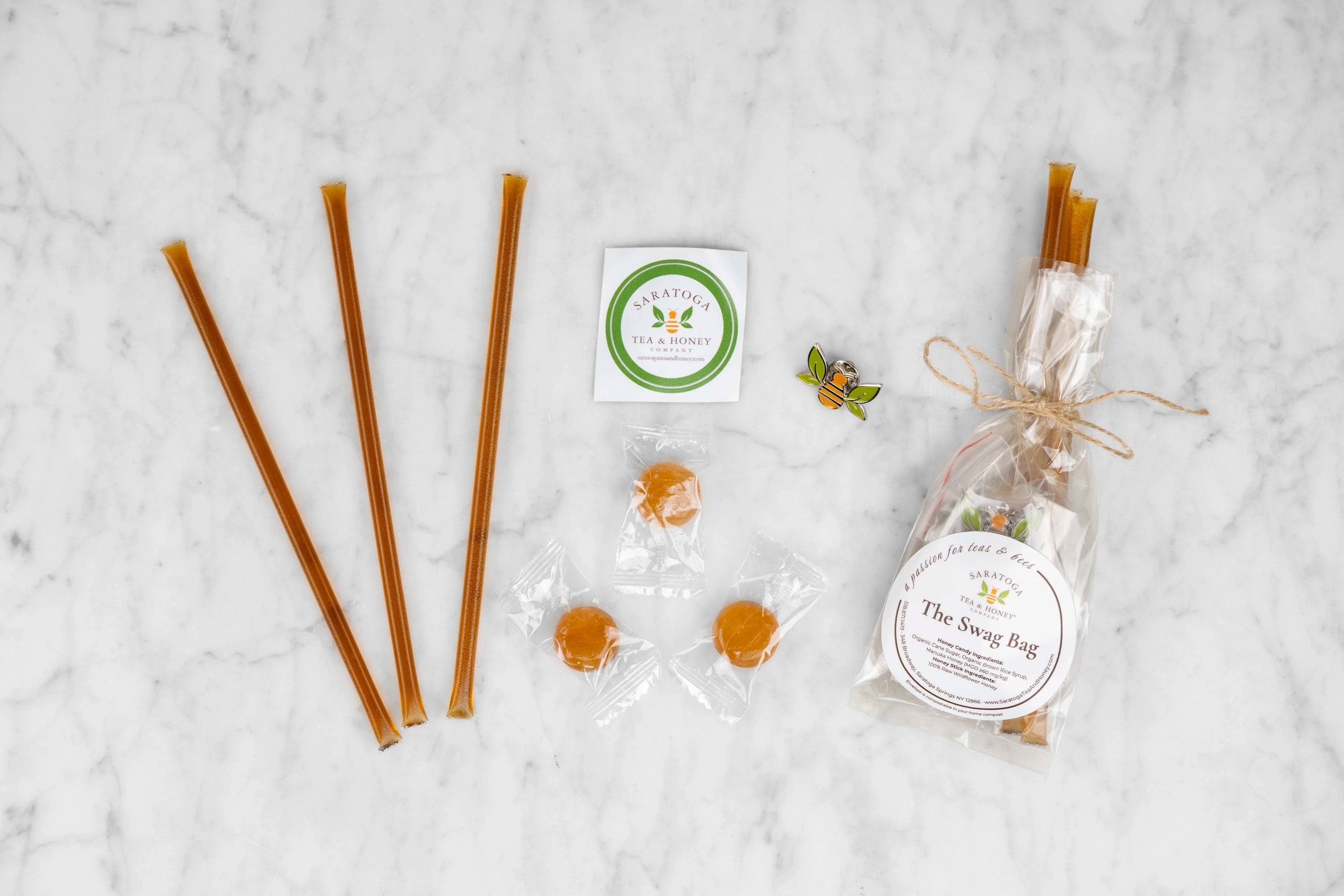 The Swag Bag contents and example product. Three honey sticks, three round hard honey candies, a Saratoga Tea & Honey Co. sticker and a tea bee enamel pin next to a composed "Swag Bag".