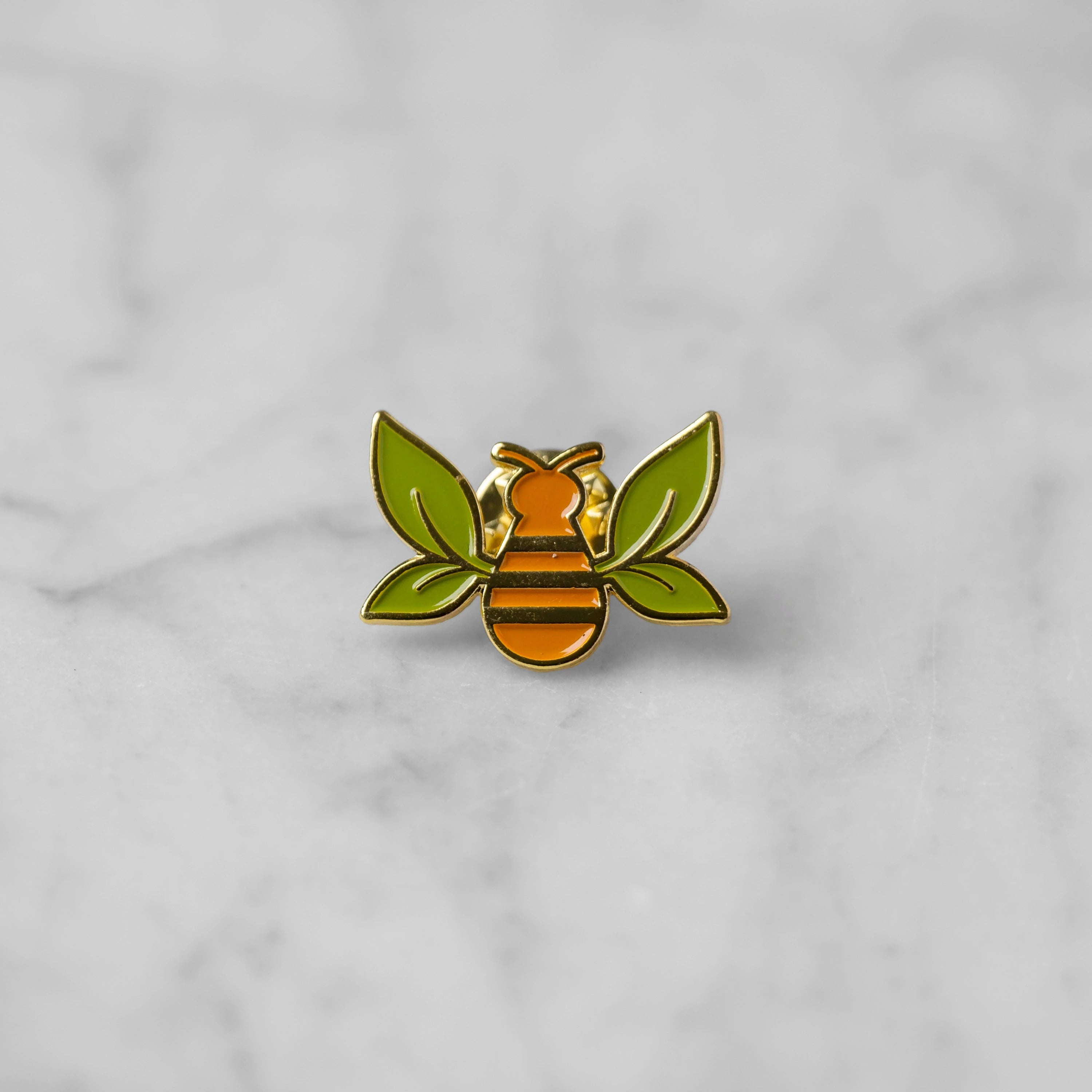 "Tea Bee" gold enamel pin with green tea leaf wings and orange bee body centered on marble. 
