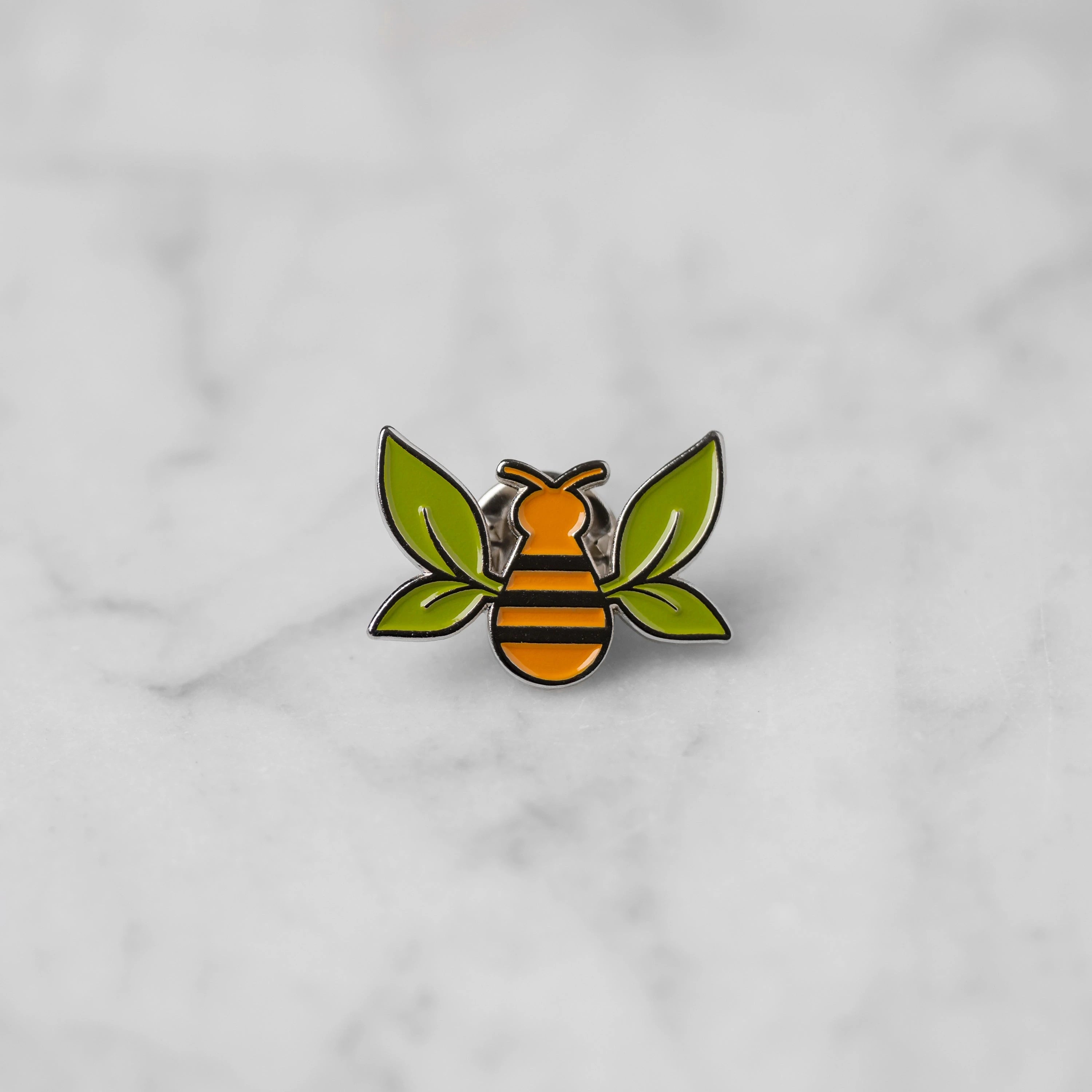 "Tea Bee" silver enamel pin with green tea leaf wings and orange bee body centered on marble. 