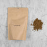 Hojicha Powder packaging and pile of ground tea. The hojicha powder comes in a kraft pouch with a label reading: Hojicha Powder 100g. Next to the pouch is a pile of ground roasted green tea. It appears brown in color and is finely ground. 