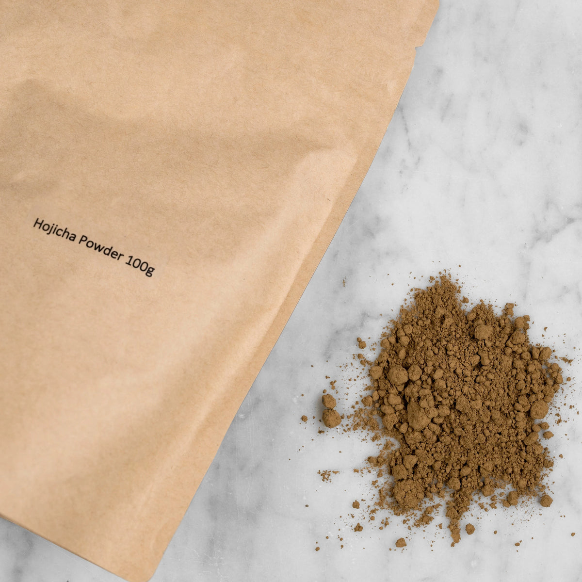 Closeup of Hojicha Powder packaging and pile of ground tea. The hojicha powder comes in a kraft pouch with a label reading: Hojicha Powder 100g. Next to the pouch is a pile of ground roasted green tea. It appears brown in color and is finely ground. 