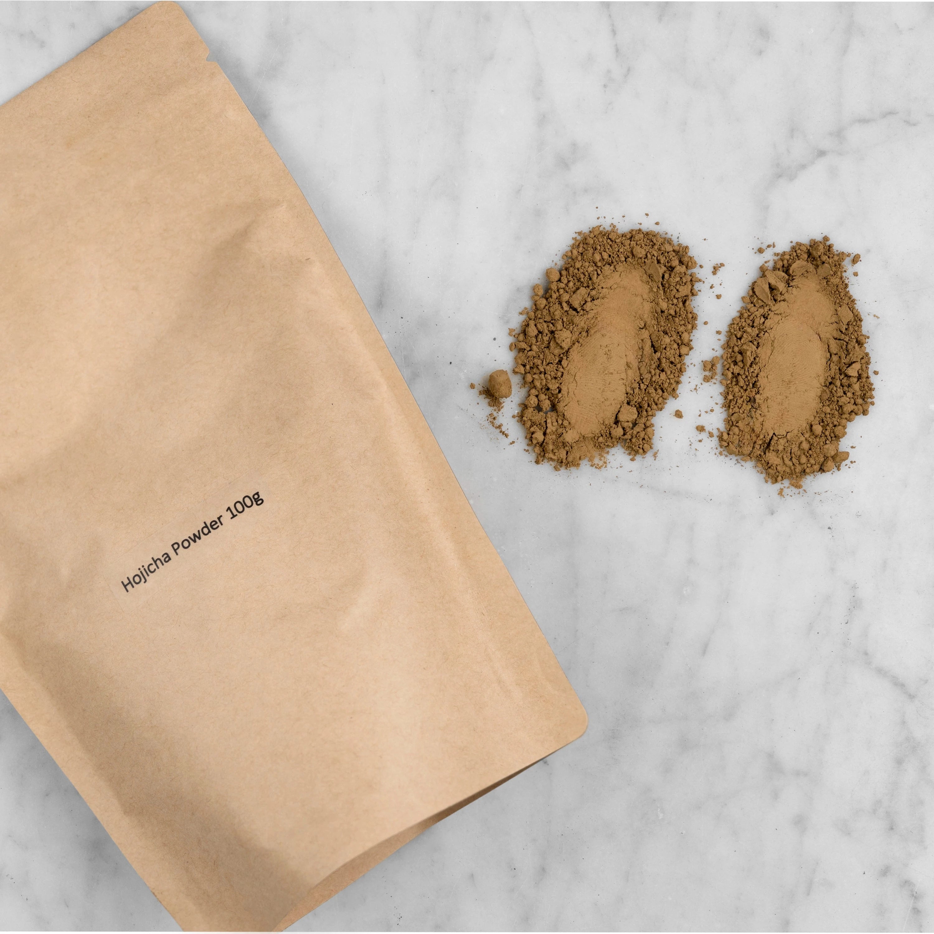 Hojicha Powder packaging and pile of ground tea. The hojicha powder comes in a kraft pouch with a label reading: Hojicha Powder 100g. Next to the pouch are two piles of hojicha powder that have been pressed and dragged to show the fine quality of the grind.