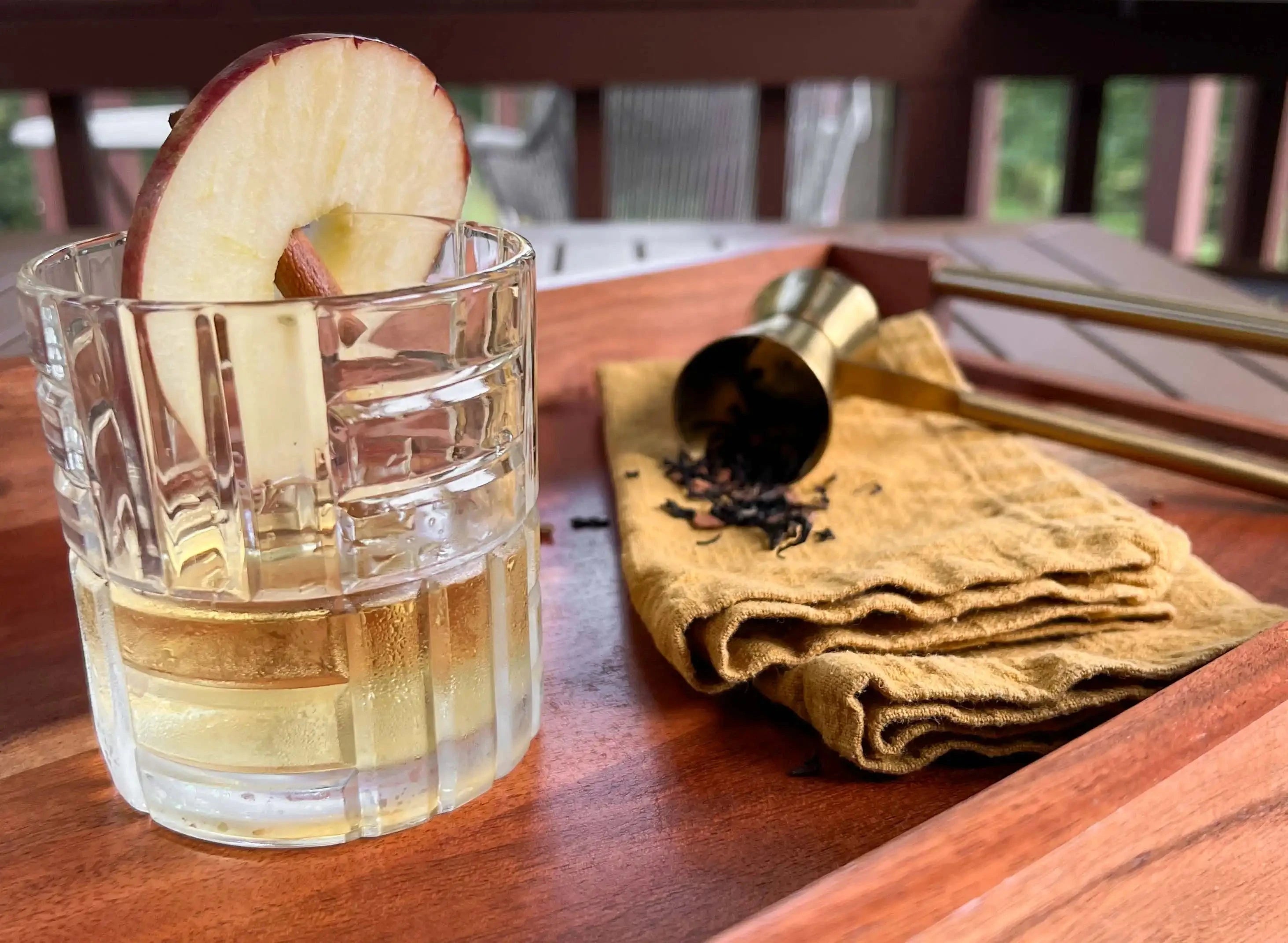 cocktail glass with tea martini, wheel of apple and cinnamon stick next to a napkin with tea spilling out of a cocktail jigger