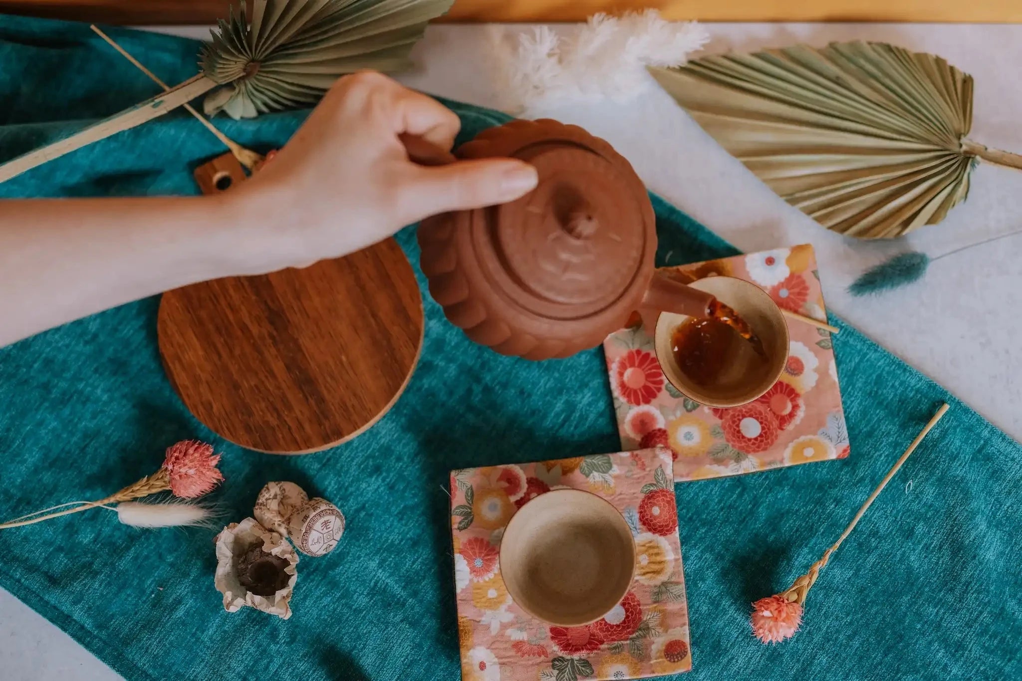 hand pouring aged tea from a yixing pot into small cups on a summery jewel toned background