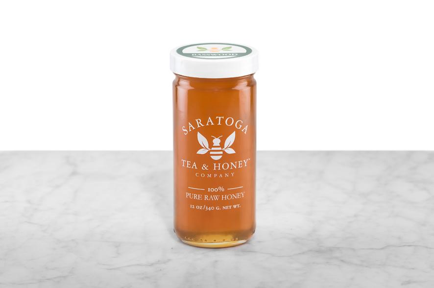 Amber-hued Basswood Honey displayed in a 12oz cylindrical glass jar with a white lid. The logo and the words: "100% Pure Raw Honey" are printed on the jar.
