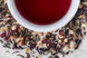 Joyful Joyful holiday tea blend leaves and brewed tea. Colorful green and black tea leaves, hibiscus flowers, and spices surround a cup of crimson-hued brewed tea.