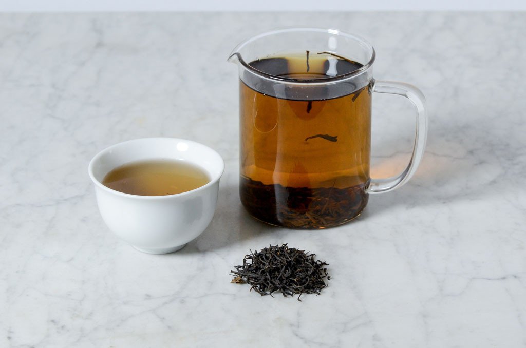 infused black tea in a white cup beside a glass infuser presented with a pile of black tea leaves on a marble background