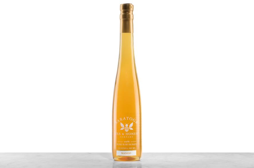 Golden Mango Infused Honey displayed in a tall, 19oz glass bottle shaped like a skinny wine bottle and capped with a cork. Bottle is printed with the logo and the words: "100% Pure Raw Honey".
