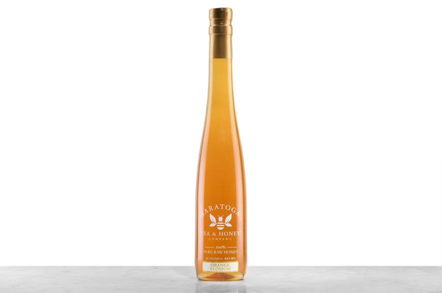 Light-amber Orange Blossom Honey displayed in a tall, 19oz glass bottle shaped like a skinny wine bottle and capped with a cork. Bottle is printed with the logo and the words: "100% Pure Raw Honey".
