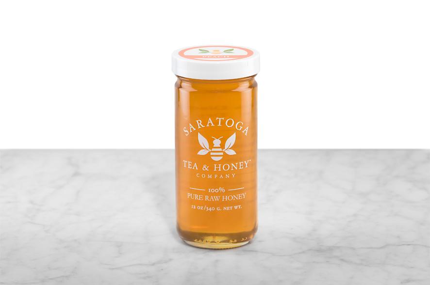 Golden-amber Peach Infused Honey displayed in a 12oz cylindrical glass jar with a white lid. The logo and the words: "100% Pure Raw Honey" are printed on the jar.
