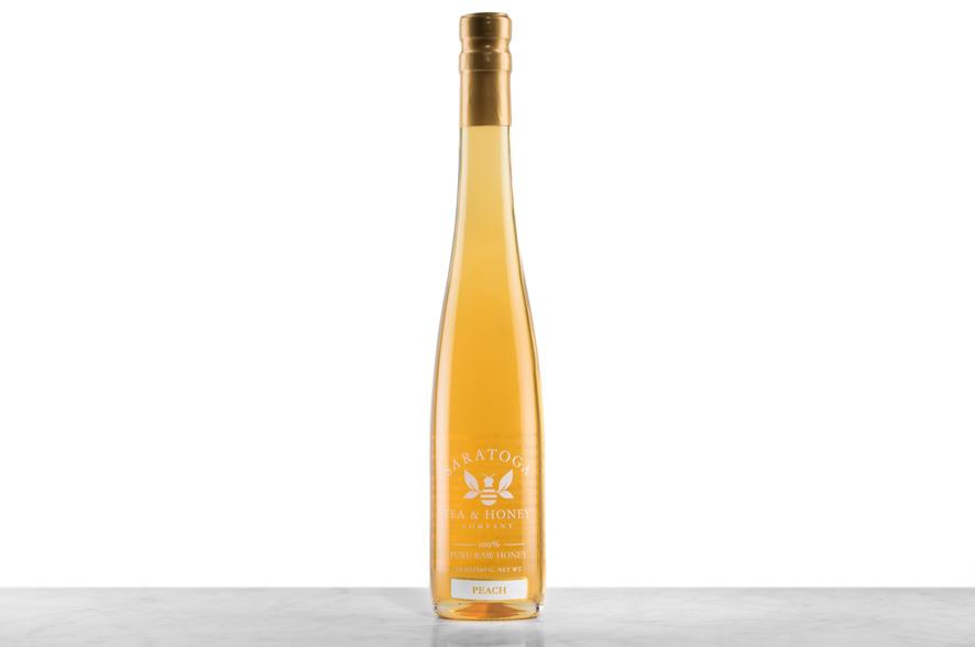 Golden-amber Peach Infused Honey displayed in a tall, 19oz glass bottle shaped like a skinny wine bottle and capped with a cork. Bottle is printed with the logo and the words: "100% Pure Raw Honey".