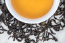 Qi Lan Wuyi oolong leaves and brewed tea. Large, dark brown twisted leaves surround a cup of amber-colored brewed tea.