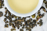 Shan Lin Xi leaves and brewed tea. Rolled, lightly oxidized oolong leaves surround a cup of pale-gold brewed tea.