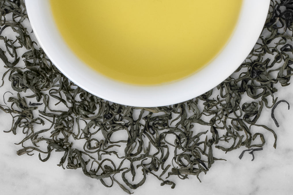 Tan Huong leaves and brewed tea. Very curly, twisted dark green tea leaves surround a cup of verdant, yellow-green brewed tea.