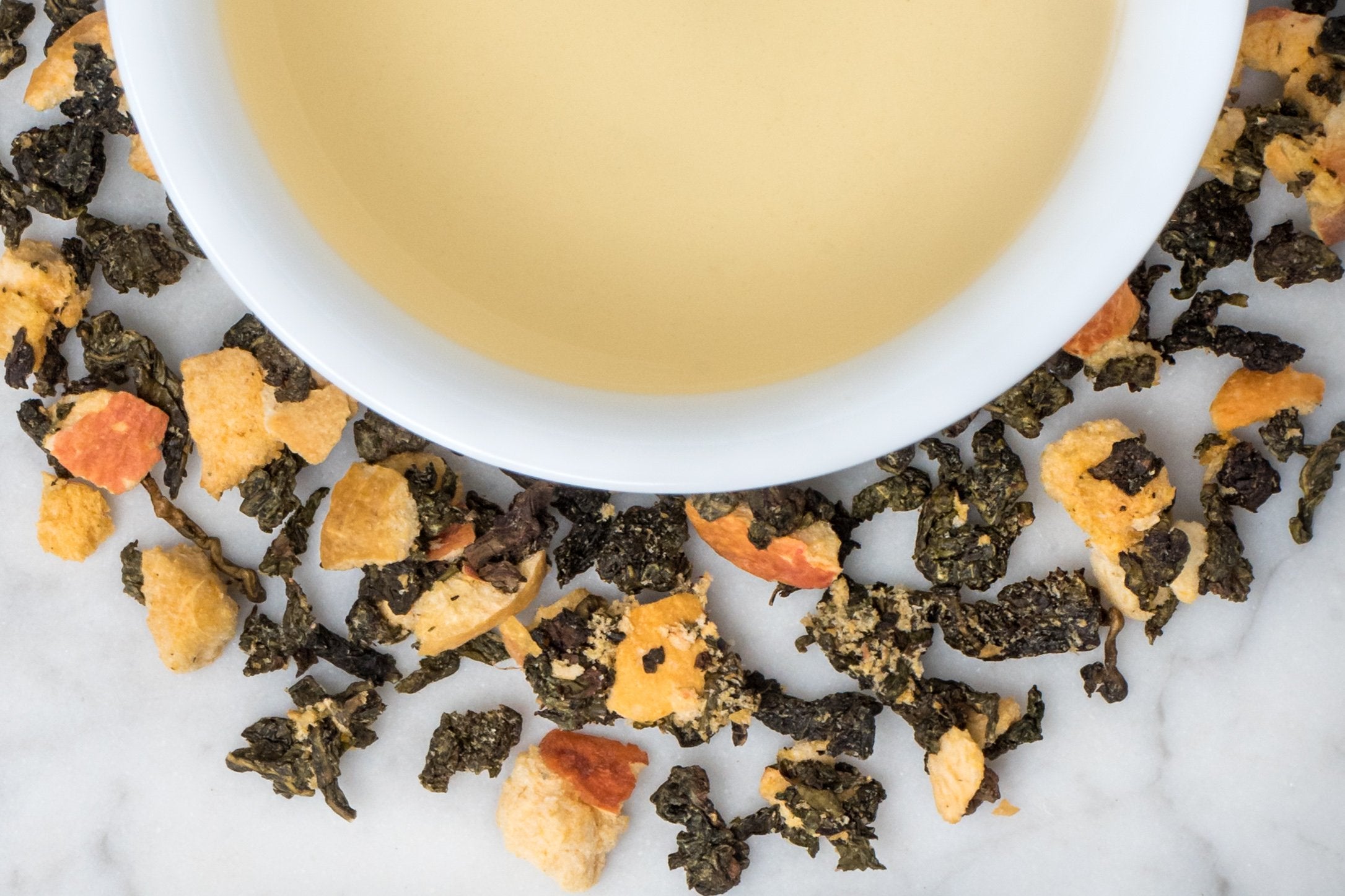 The Apricot Queen leaves and brewed tea. Rolled oolong and dried apricot bits surround a cup of pale yellow brewed tea.