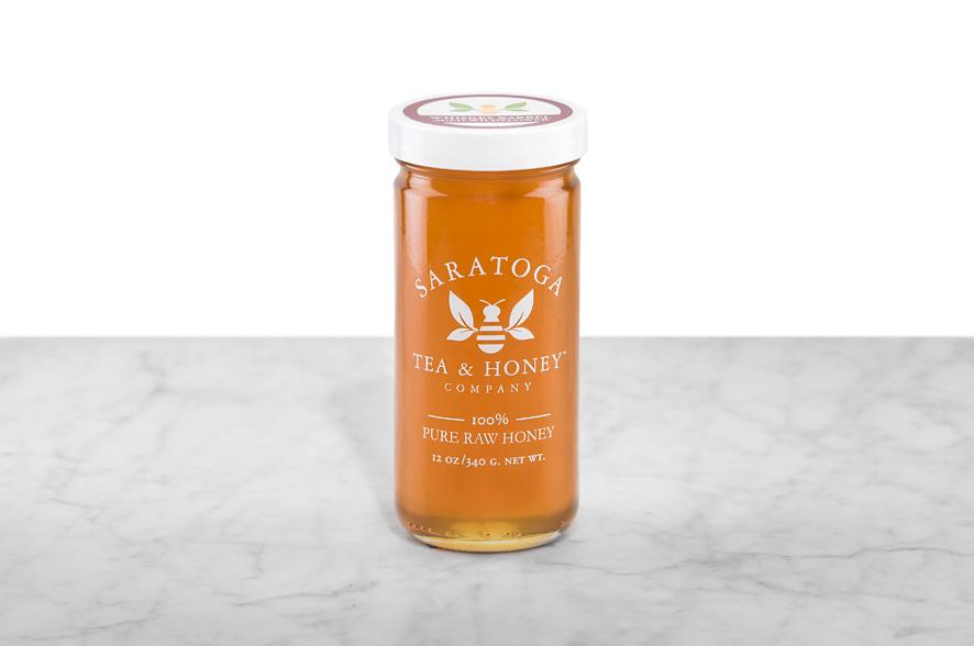 Amber-hued Saratoga Springs Whiskey Barrel Aged Wildflower Honey displayed in a 12oz cylindrical glass jar with a white lid. The logo and the words: "100% Pure Raw Honey" are printed on the jar.