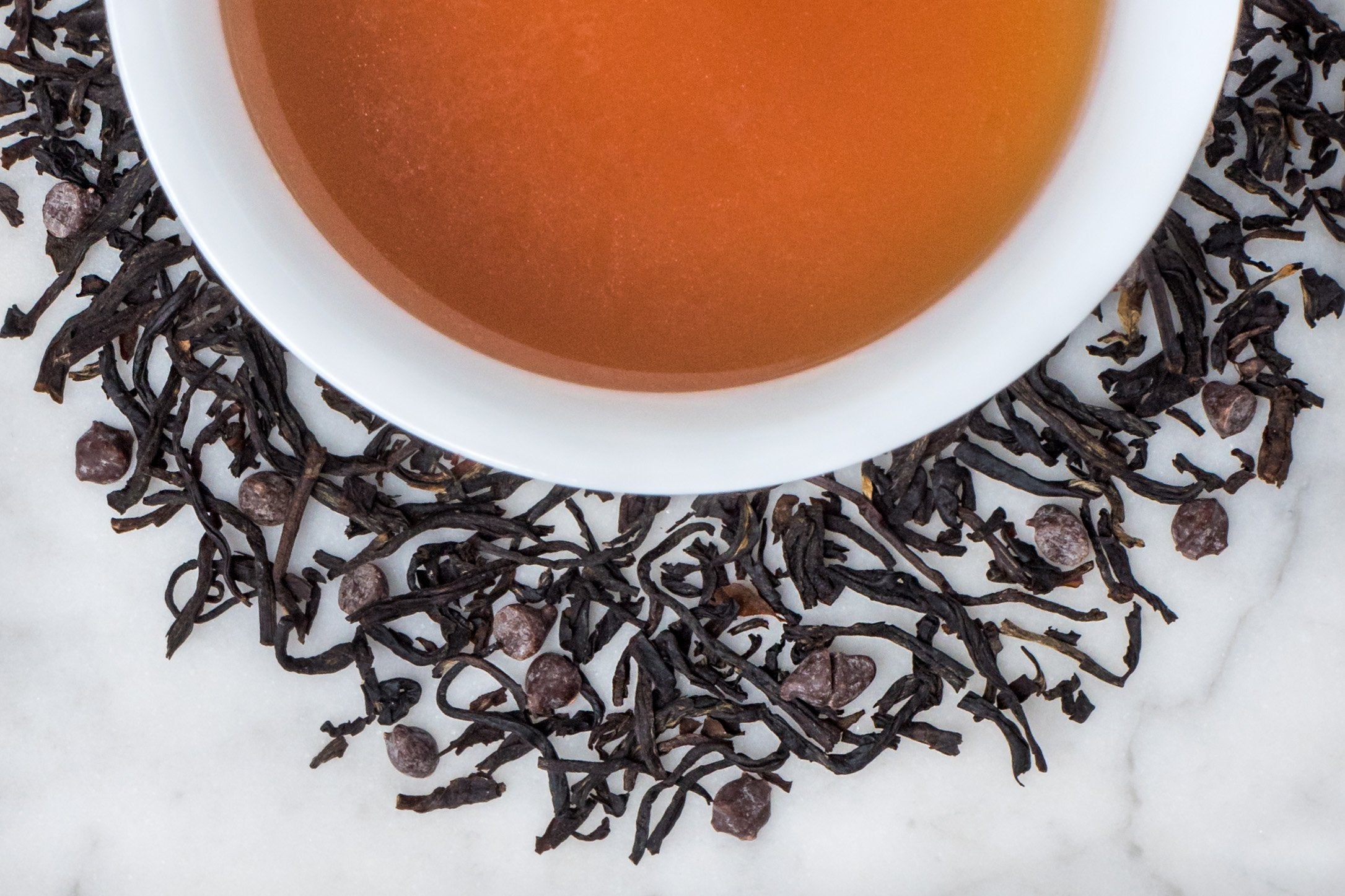 Coco Shou leaves and brewed tea. Long, twisted black tea leaves and tiny chocolate kisses surround a cup of dark-brown brewed tea.