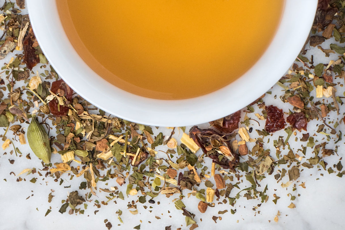 Exhale leaves and brewed tea. Small leaves, berries, and cardamom pods surround a cup of amber-hued brewed herbal tea.