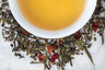 Green Paddock Pom Pom leaves and brewed tea. Black and green tea leaves and dried fruit surround a cup of light gold-colored brewed tea.