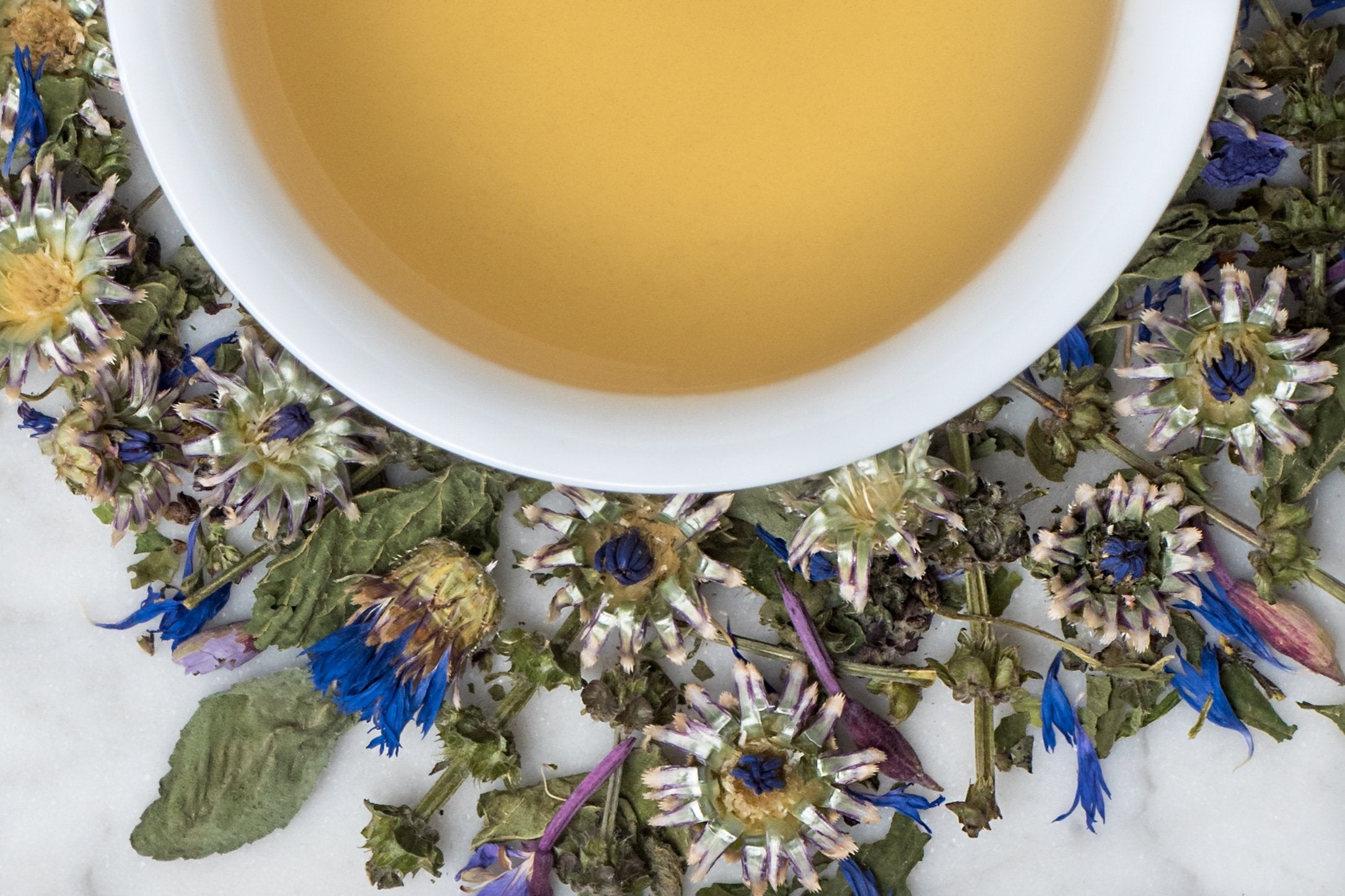 Tulsi Supreme leaves and brewed tea. Green leaves and blue flowers surround a cup of brewed, light khaki-colored herbal tea.
