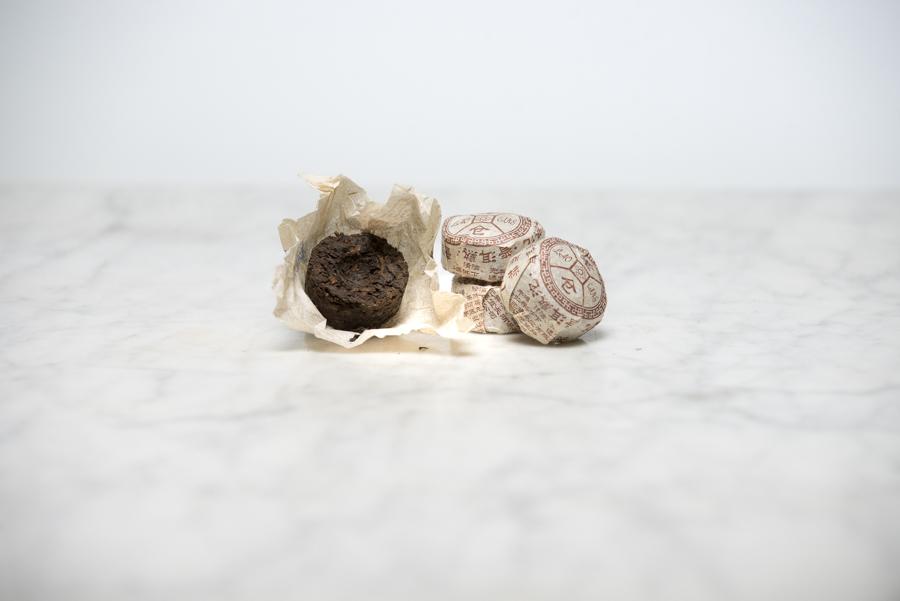 2012 Menghai tea packaging. An unwrapped toucha (small aged tea cake) leans on three small, wrapped tea nests of 2012 Menghai pu erh. 