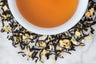 One Night in Rio leaves and brewed tea. Twisted black tea leaves with coconut and pineapple pieces surrounds a cup of medium-brown brewed tea.
