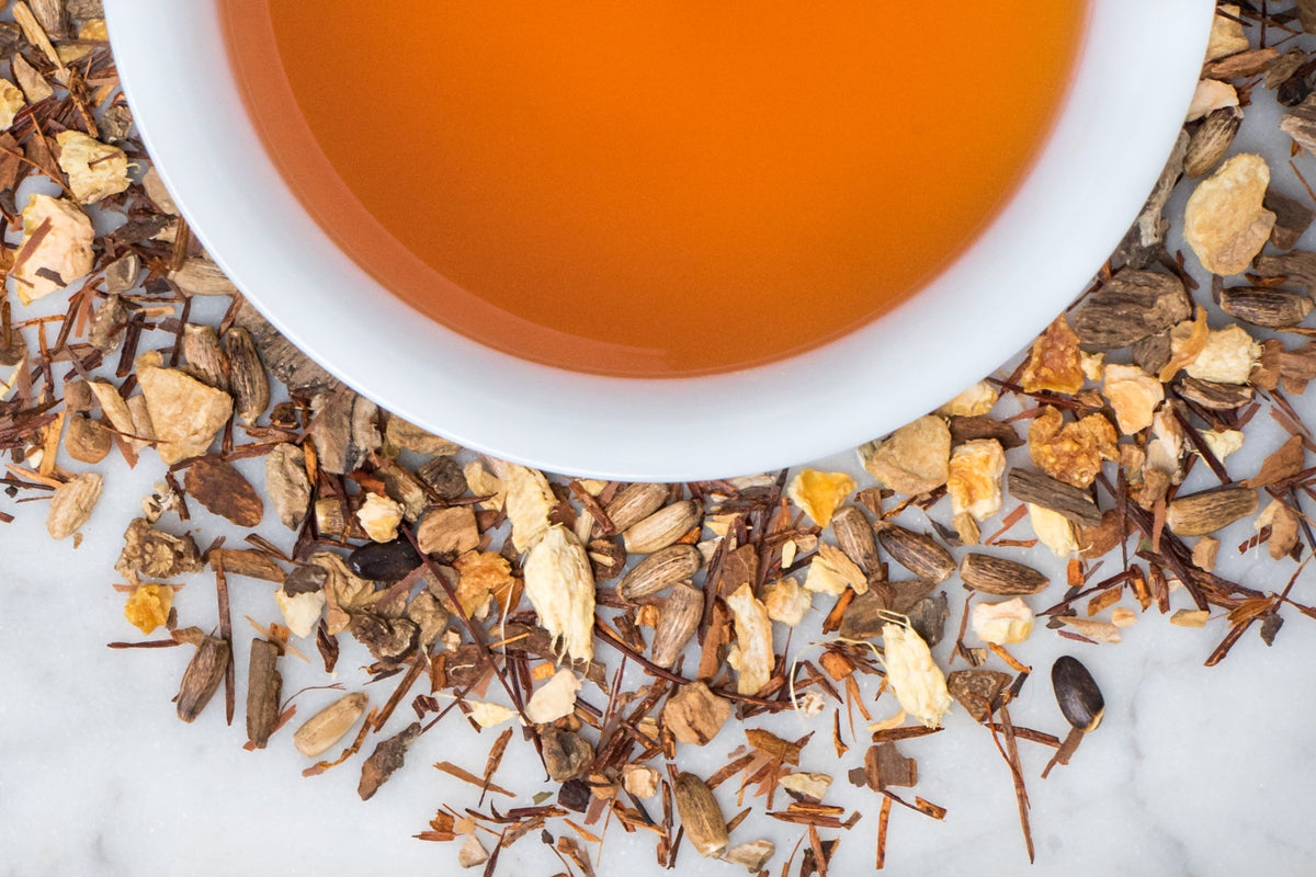Teatoxify leaves and brewed tea. A granola-like collection of roots and leaves surrounds a cup of russet-hued brewed herbal tea.