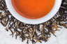 Yunnan Da Ye leaves and brewed tea. Twisted dark brown leaves and golden brown buds surround a cup of reddish-brown brewed black tea.